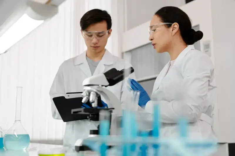 Two scientists in lab coats and goggles discussing results beside a microscope