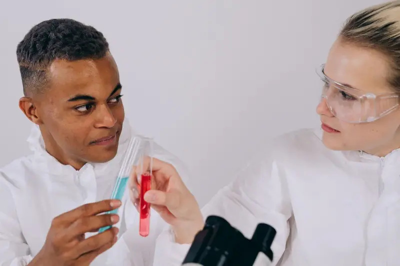 Two young scientists in lab coats and goggles comparing sample tubes and smiling