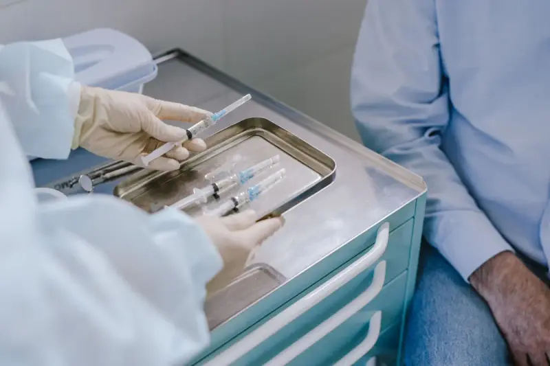 Gloved hand lifting a prepared syringe from a steel tray on a medication cart