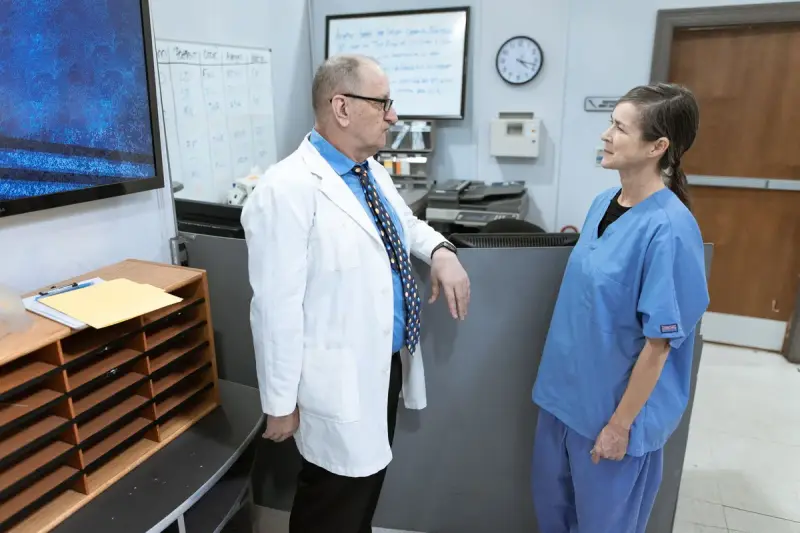 A clinician in a white coat talks with a nurse in blue scrubs at a hospital unit desk