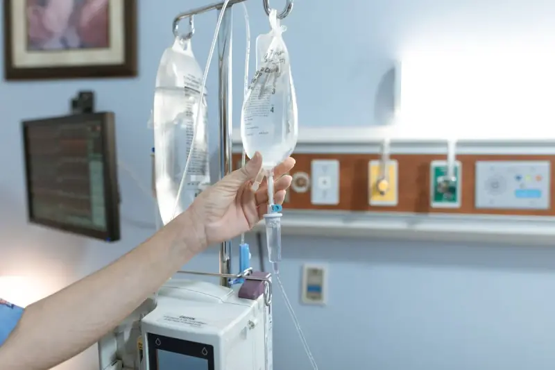 Clinician's hand checking IV bags on a pole above an infusion pump in a hospital room