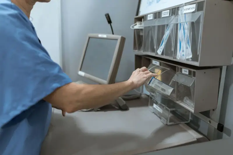 Hand pulling a supply from a lit drawer of an automated medication cabinet beside a touchscreen