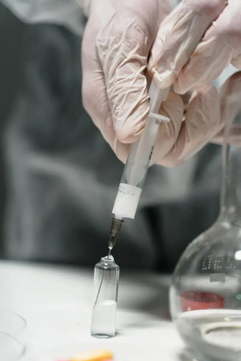 Gloved hands inserting a syringe needle into a small glass vial beside a flask