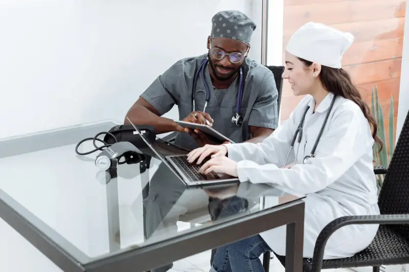 Two clinicians at a glass table working on a laptop and a tablet together