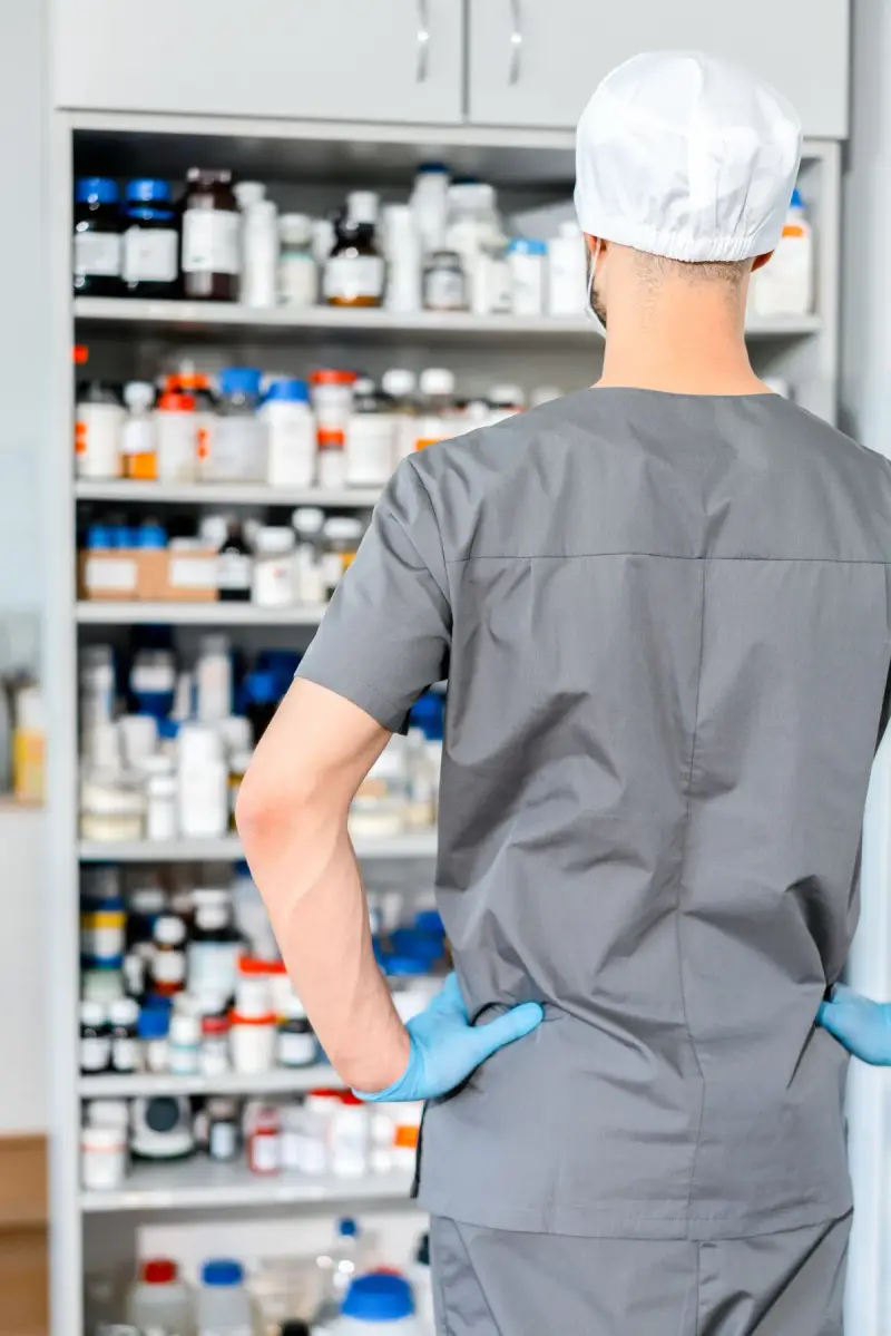 Pharmacist in grey scrubs, cap and blue gloves facing shelves of medicine bottles