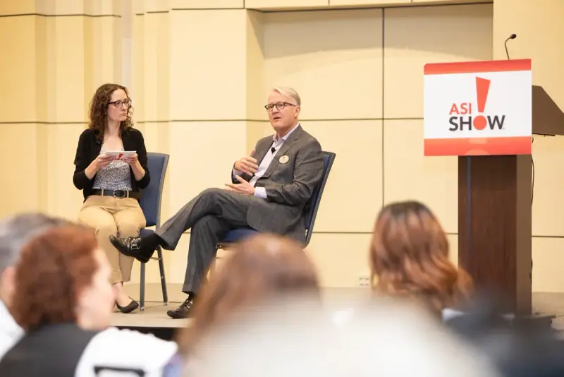 Two speakers seated on stage for a panel next to an ASI Show lectern