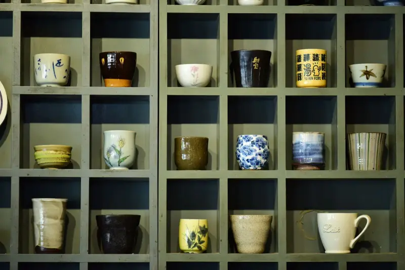 Assorted ceramic mugs and cups arranged in a grid of green wooden cubbies