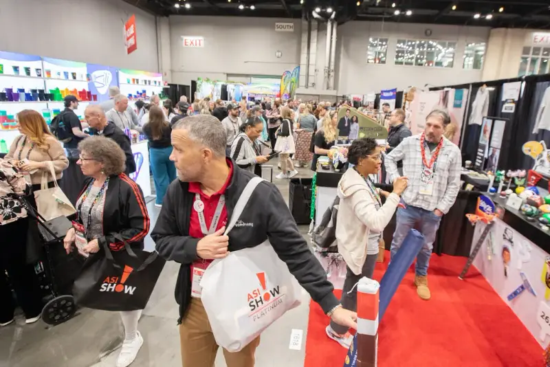 Attendees carrying show tote bags along a busy aisle of the ASI Show Orlando exhibit hall