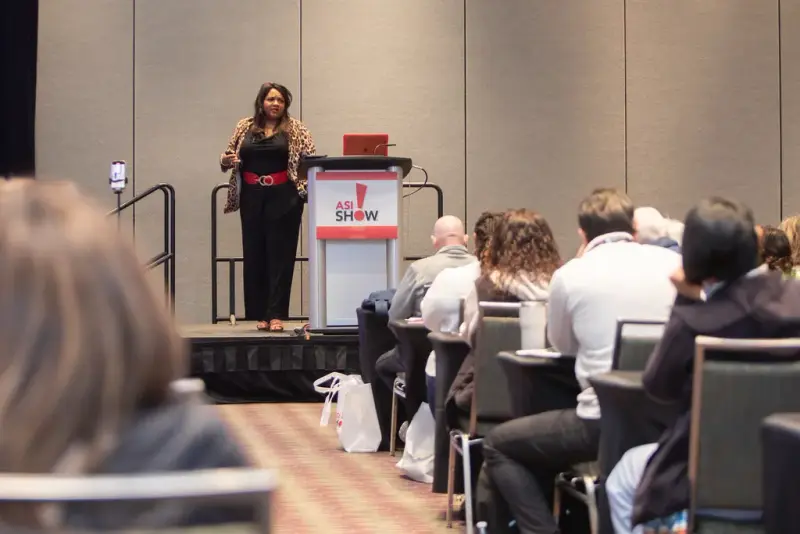 Presenter on stage beside an ASI Show lectern speaking to a seated audience