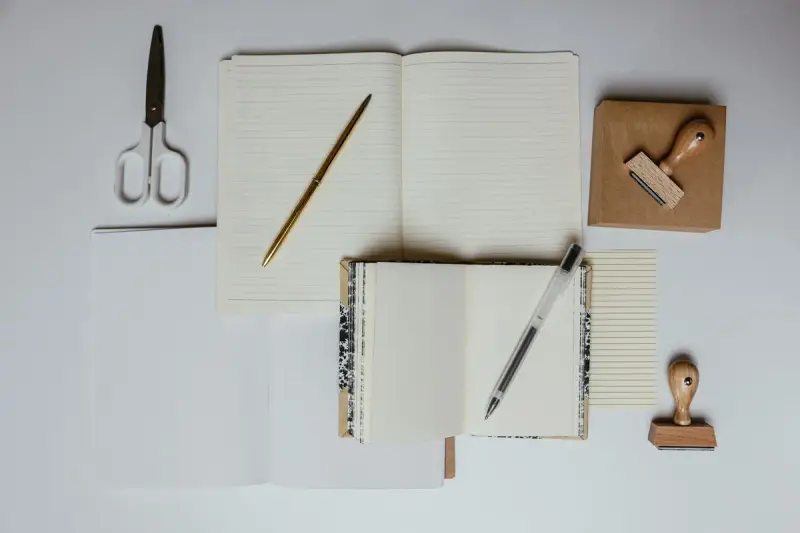 Flat lay of open notebooks, pens, scissors and rubber stamps on a light desk