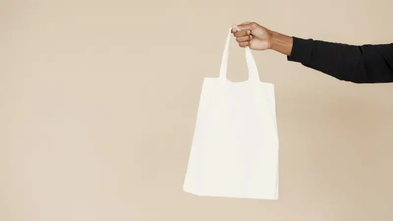 Hand holding a plain white canvas tote bag against a beige background