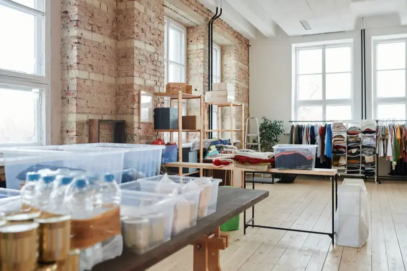 Bright brick-walled stockroom with bins, boxes and racks of clothing by tall windows