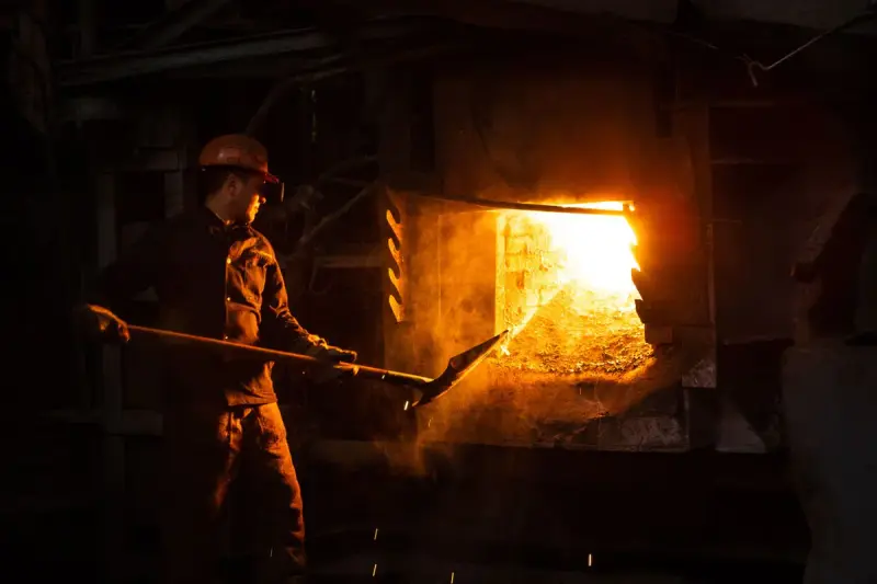 Worker in a hard hat tending an open industrial furnace glowing orange