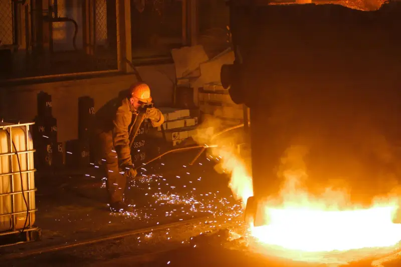 Steelworker in a helmet and mask working beside a glowing pool of metal as sparks fly