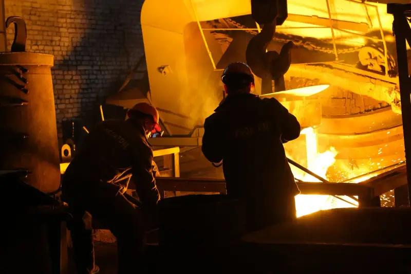 Two workers in hard hats tending glowing molten metal at a furnace
