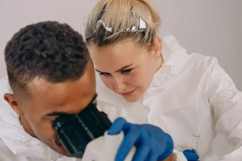 Two scientists in lab coats and protective gear working at a microscope