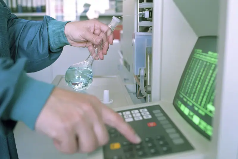 Scientist holding a glass flask while entering settings on an analytical instrument keypad