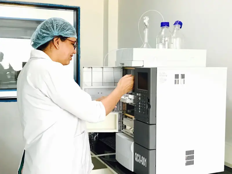 Female scientist in a lab coat and hair cover operating a liquid chromatography system