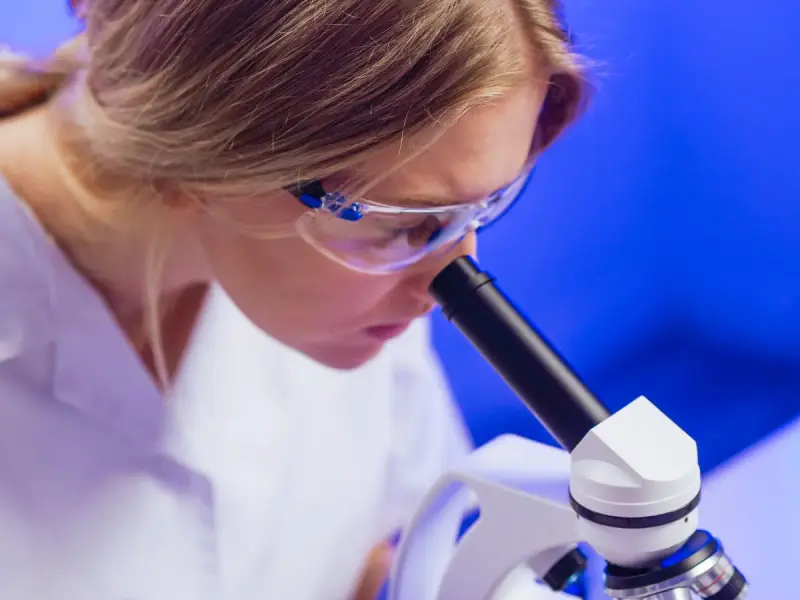 Woman scientist in a white coat and safety glasses looking into a microscope