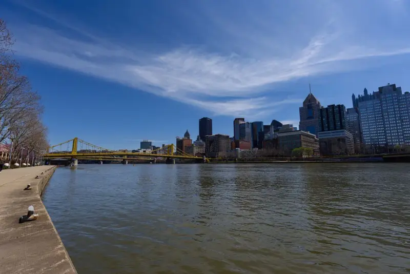 Pittsburgh skyline across the river with a yellow suspension bridge under blue sky