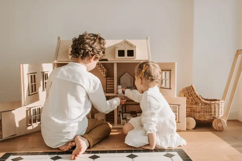 Two young children seen from behind playing with a large wooden dollhouse in a sunny room