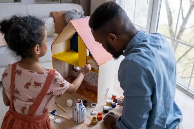 Father and young daughter painting a wooden dollhouse together by a bright window
