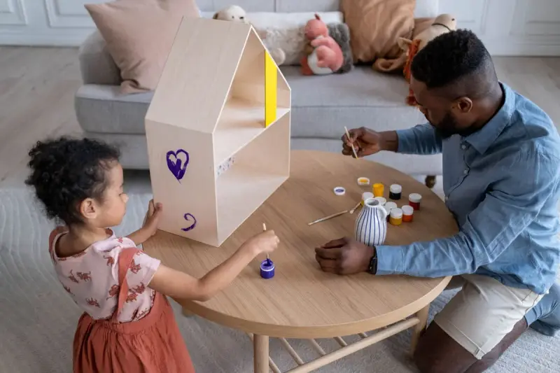 Father kneeling at a low table painting with his daughter beside a wooden toy house