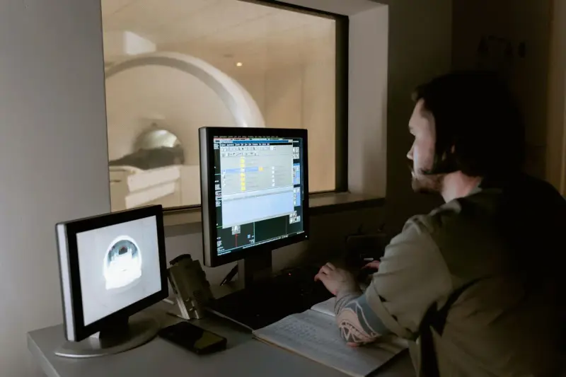 Technologist at a control console watching monitors, with a scanner room visible through the window