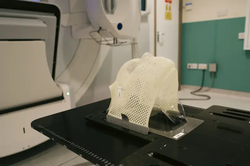 White mesh immobilization mask mounted on a carbon-fiber treatment couch beside a radiotherapy machine