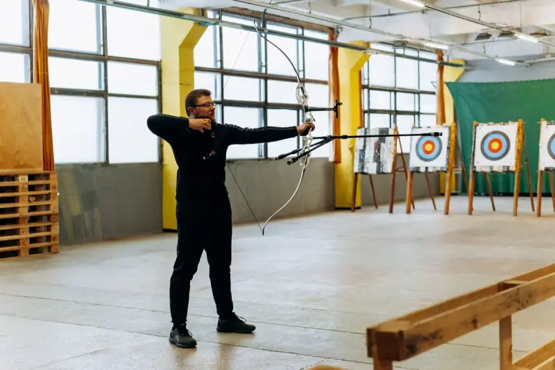 Archer drawing a bow toward targets across a bright indoor archery range