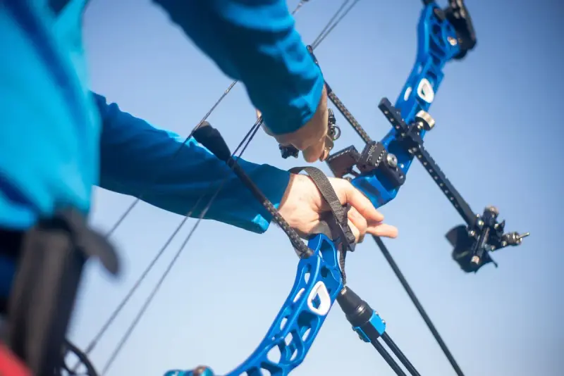 Hands gripping a blue compound bow with sight and stabilizer against the sky