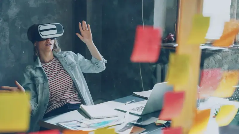 Woman using a virtual reality headset at a desk with a laptop and notes
