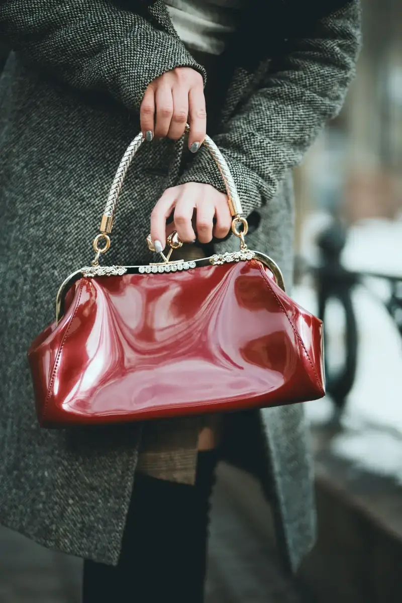 Hands holding a glossy oxblood frame handbag against a gray wool coat