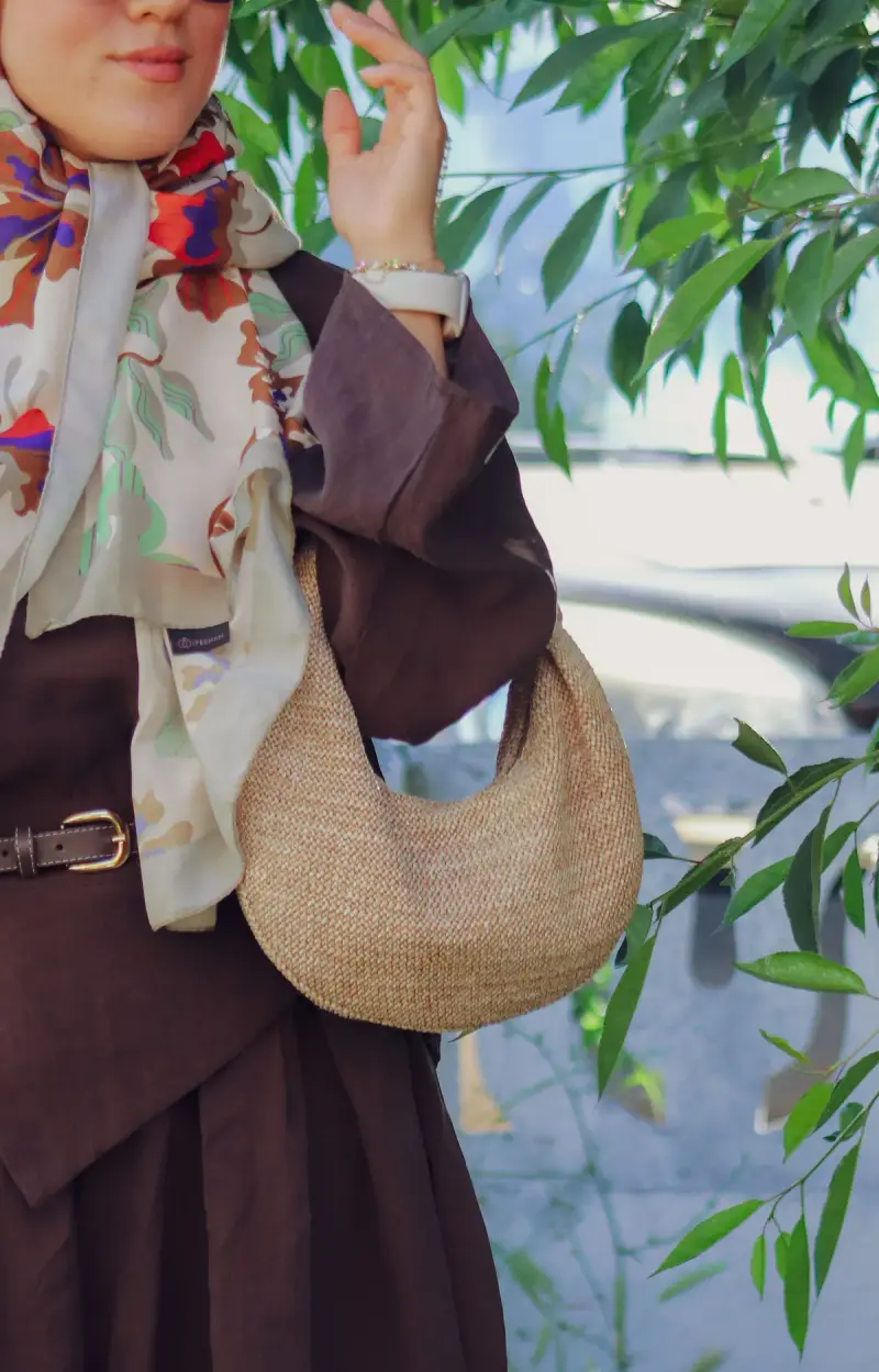 Woman in a printed silk scarf and brown coat carrying a woven straw crescent bag