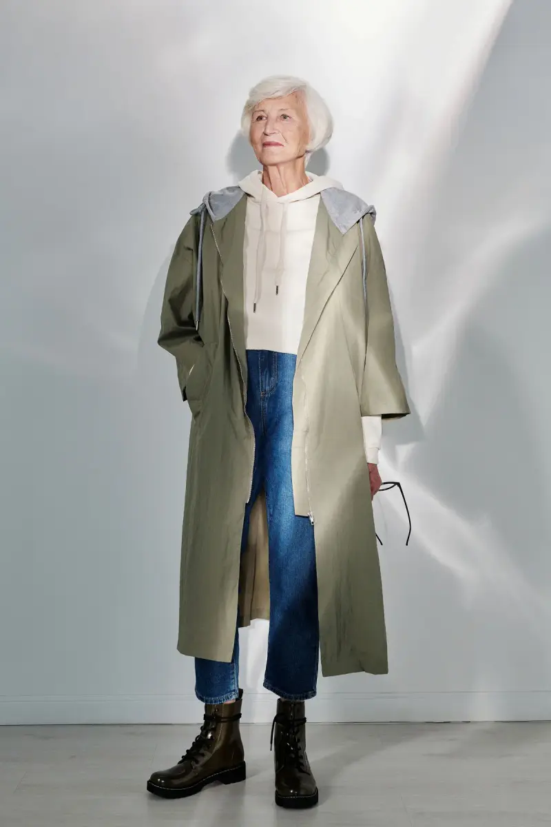 Silver-haired woman in a long olive trench, white hoodie, jeans and combat boots
