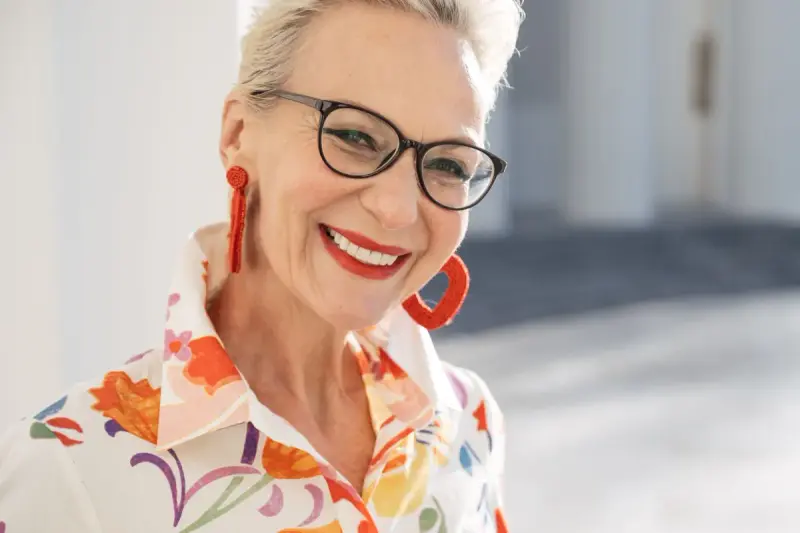 Smiling silver-haired woman in black glasses, red hoop earrings and a floral shirt