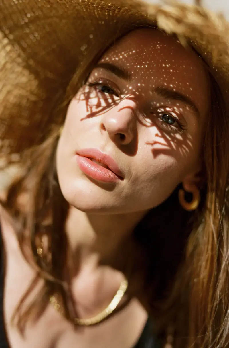 Woman in a straw hat with dappled sunlight on her face, wearing gold hoops and a gold chain