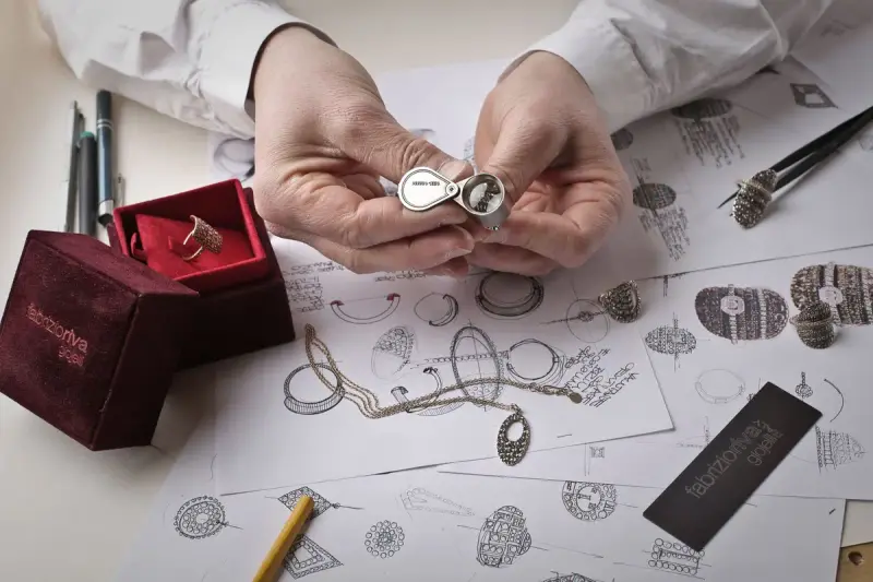Hands inspecting a diamond ring with a loupe over a bench covered in sketches