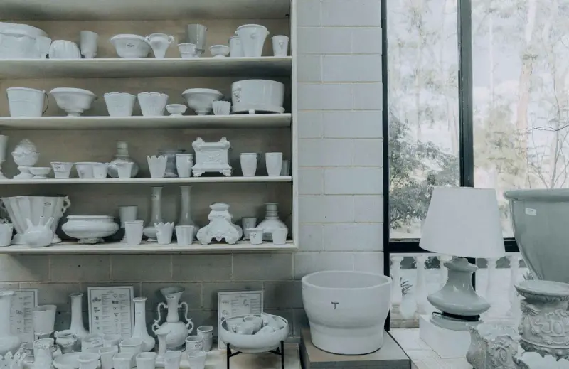 White shelves filled with unglazed ceramic bowls, vases and lamps in a bright workshop