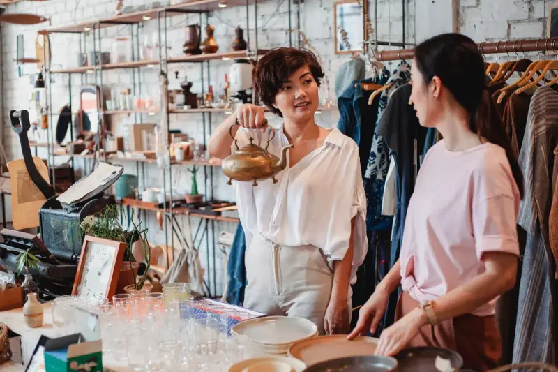 Woman holding up a brass kettle to show a friend in a busy homewares and clothing shop