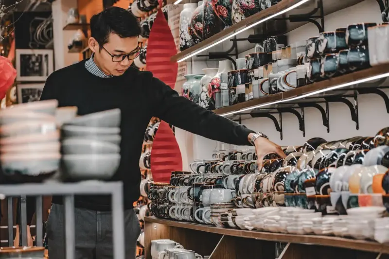 Man browsing shelves of patterned ceramic mugs and stacked bowls in a pottery shop