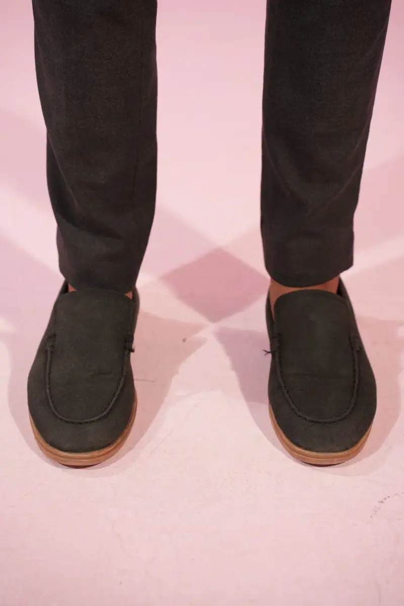 Dark suede loafers with tan soles worn with charcoal trousers on a pink backdrop