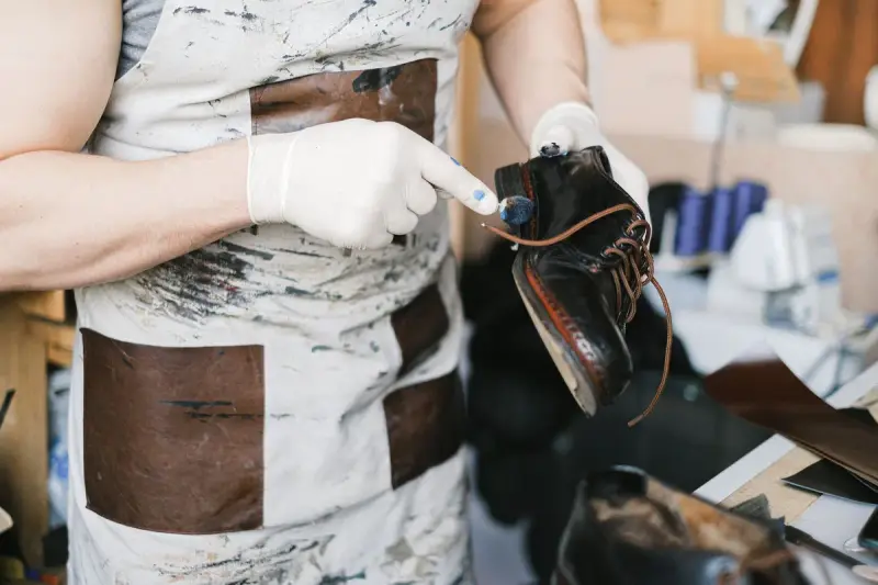 Gloved craftsman in a work apron checking a black leather shoe in a workshop