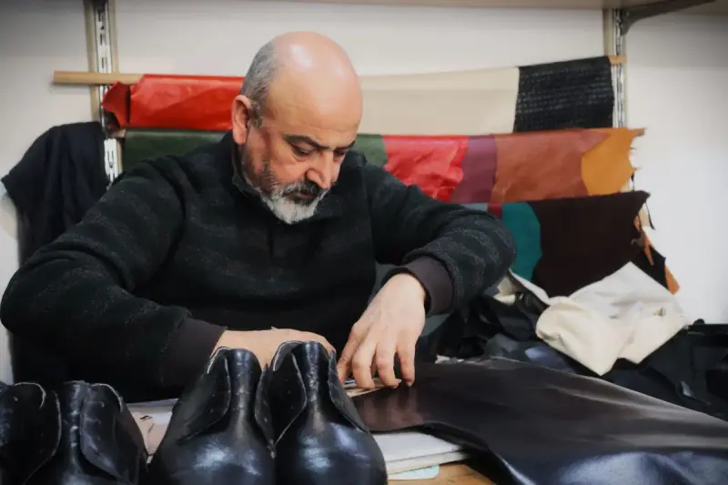 Shoemaker cutting leather at a table with finished black shoes in front of him