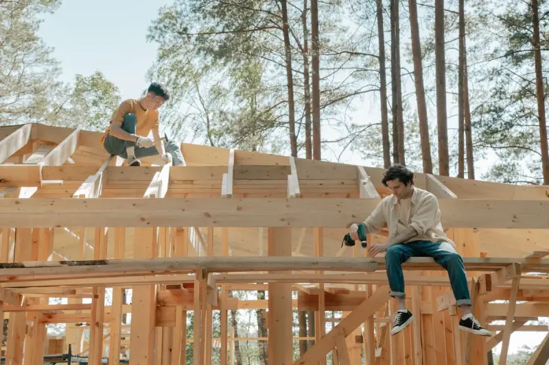 Two carpenters sitting on wooden roof trusses, one with a cordless drill, among pine trees
