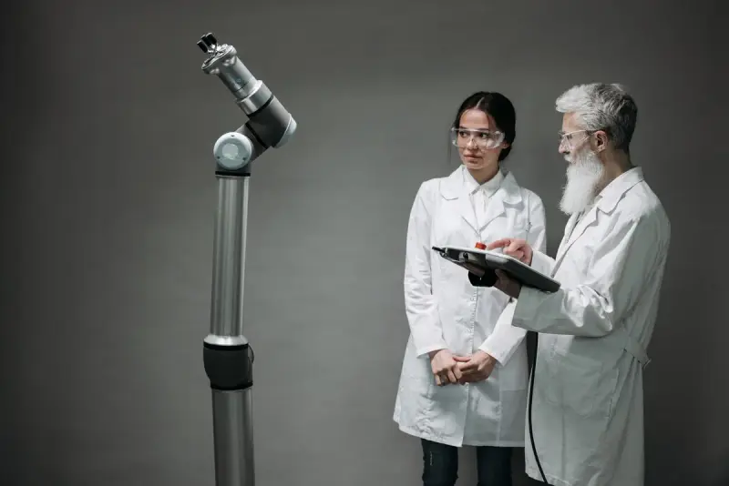 Two engineers in lab coats and safety glasses reviewing a teach pendant beside a cobot arm