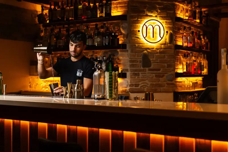 Bartender pouring a cocktail behind a backlit bar counter lined with spirit bottles