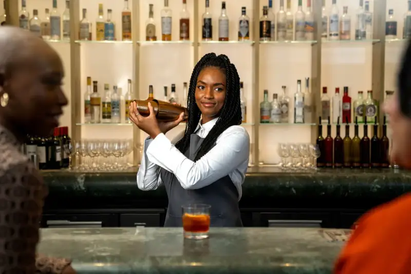 Bartender in a gray apron shaking a cocktail in front of lit bottle shelves