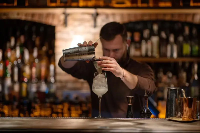 Bartender straining a cocktail into a wine glass at a warmly lit brick bar