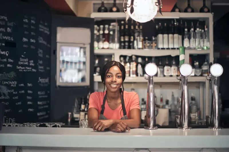 Smiling bartender in an apron leaning on a bar counter beside beer taps